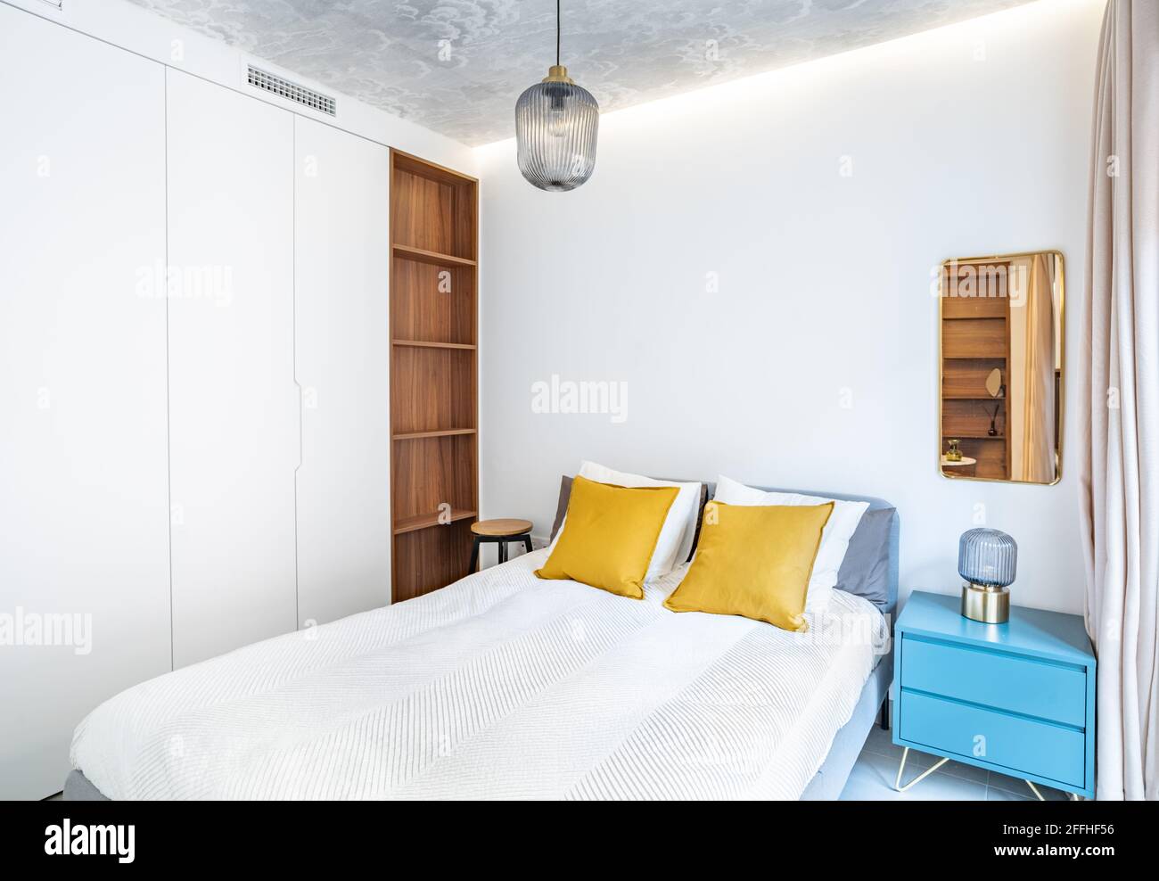 Sauberes, helles Schlafzimmer mit Bett, Bettdecke, Beistelltisch, Lampen, Vorhang, Metallspiegel, LED-indirektes Licht und bemalter Tapete an der Decke. Minimalistischer Weißkauch Stockfoto