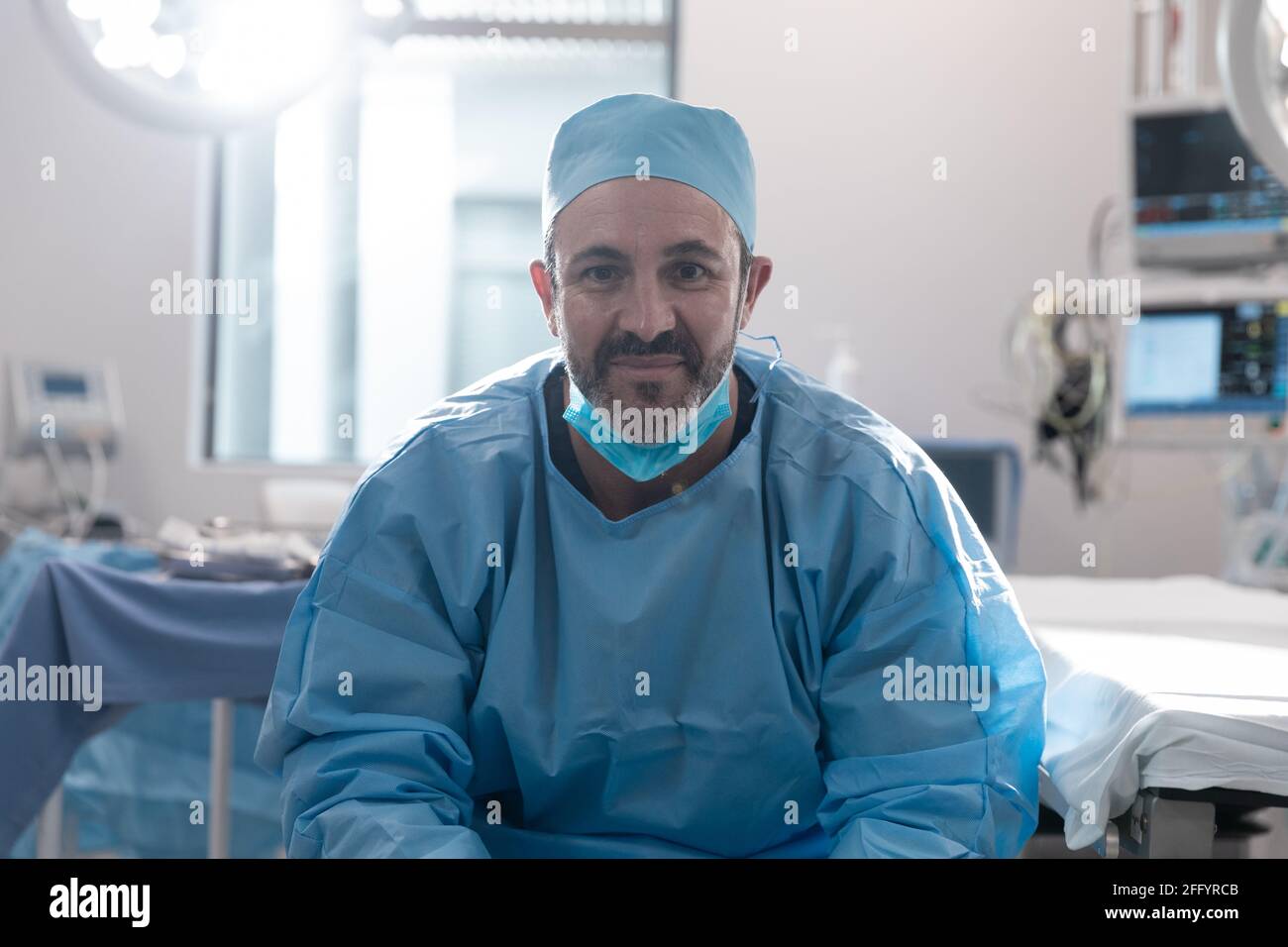 Lächelnder kaukasischer Chirurg mit Gesichtsmaske und Schutzkleidung Im Operationssaal Stockfoto