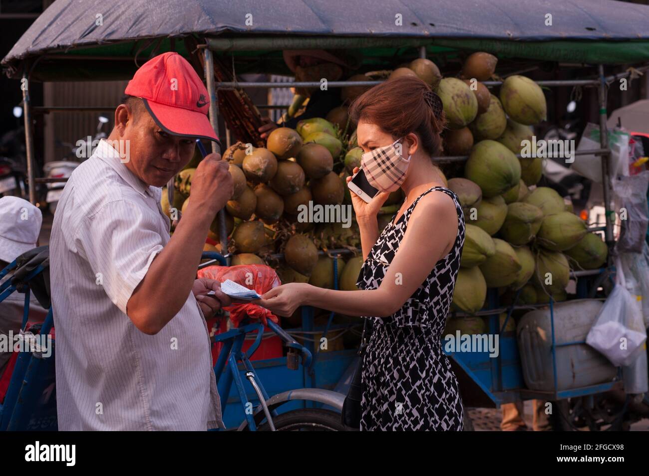 Kokosnusslieferung, kambodschanische Frau trägt eine schützende Gesichtsmaske und benutzt ein Smartphone auf dem Kandal Markt während der Coronavirus-Pandemie. Phnom Penh, Kambodscha. März 2020. © Kraig Lieb Stockfoto