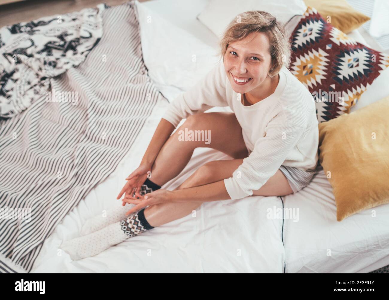 Junge blonde Frau in Pyjama gekleidet sitzen im gemütlichen Bett mit Kissen und aufrichtig lächelnd und Blick auf die Kamera. Wohnen zu Hause und süß gemütlich Stockfoto
