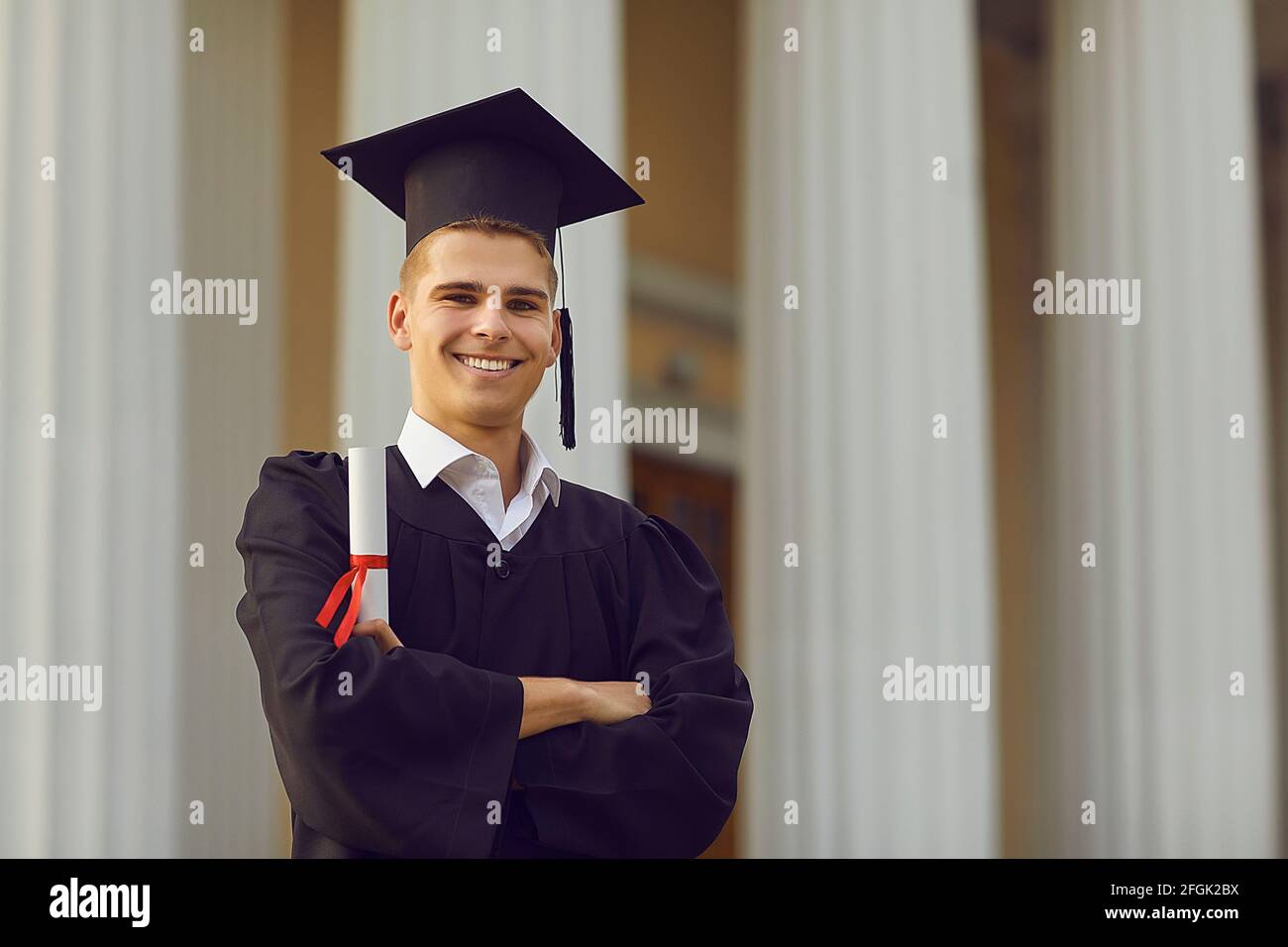 Erfolgreicher Absolvent im akademischen Kleid posiert mit Diplom in den Händen vor Hochschulhintergrund. Stockfoto