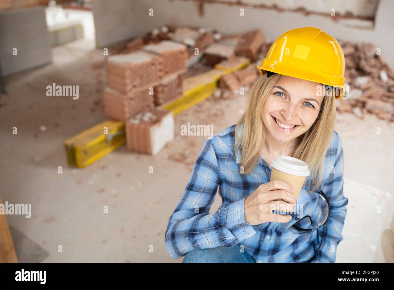 Hübsche junge Arbeiterin mit gelbem Schutzhelm nimmt Kaffee Pause auf Baustelle innen und ist glücklich Stockfoto