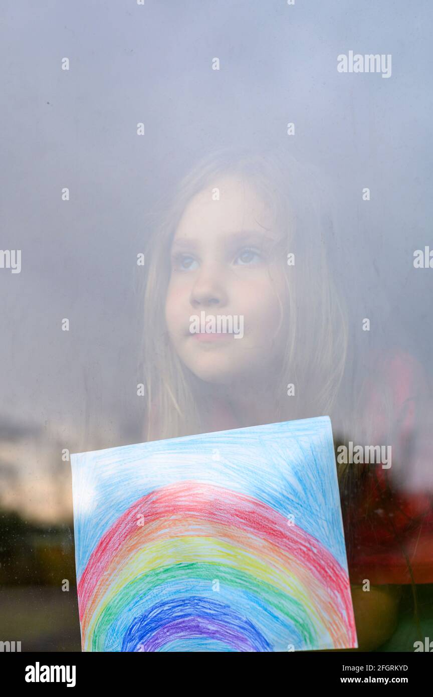 Laute Wirkung. Kind Mädchen sieben Jahre alt mit Zeichnung Regenbogen schaut durch das Fenster während covid-19 Quarantäne. Zu Hause bleiben, lassen Sie uns alle gut sein. verti Stockfoto