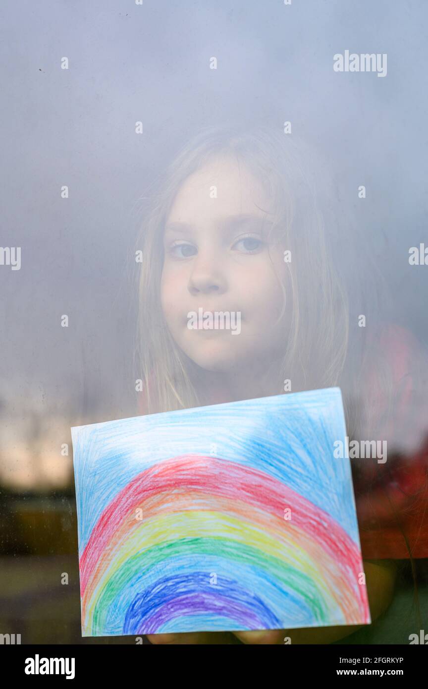 Laute Wirkung. Kind Mädchen sieben Jahre alt mit Zeichnung Regenbogen schaut durch das Fenster während covid-19 Quarantäne. Zu Hause bleiben, lassen Sie uns alle gut sein. verti Stockfoto