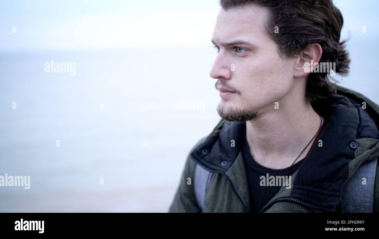 Attraktiver, blauäugiger junger Mann mit Bart und lockigem Haar Einzelporträt an der Küste Stockfoto