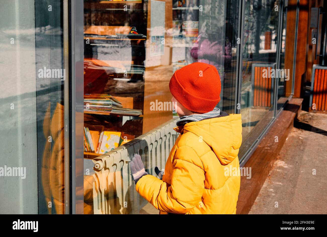Ein Kind in einer gelben Jacke untersucht aufmerksam das Fenster eines Straßenkiosks. Stockfoto