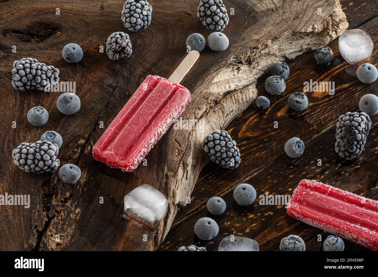 Gefrorene Brombeeren, Heidelbeeren, Eislollies und Eiswürfel über einem Holzbrett Stockfoto