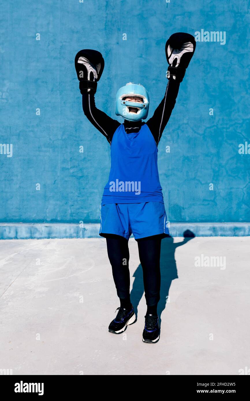 In voller Länge lächelnde reife Frau in Sportswear und Boxhandschuhen Stehen mit Helm gegen blaue Wand mit erhobenen Armen Stockfoto