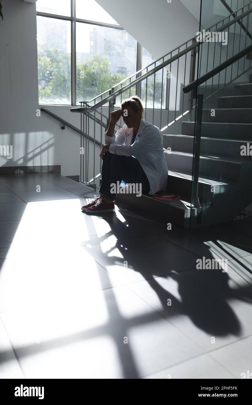 Kaukasische Ärztin, die auf der sonnigen Spitaltreppe sitzt und sich Sorgen macht Stockfoto