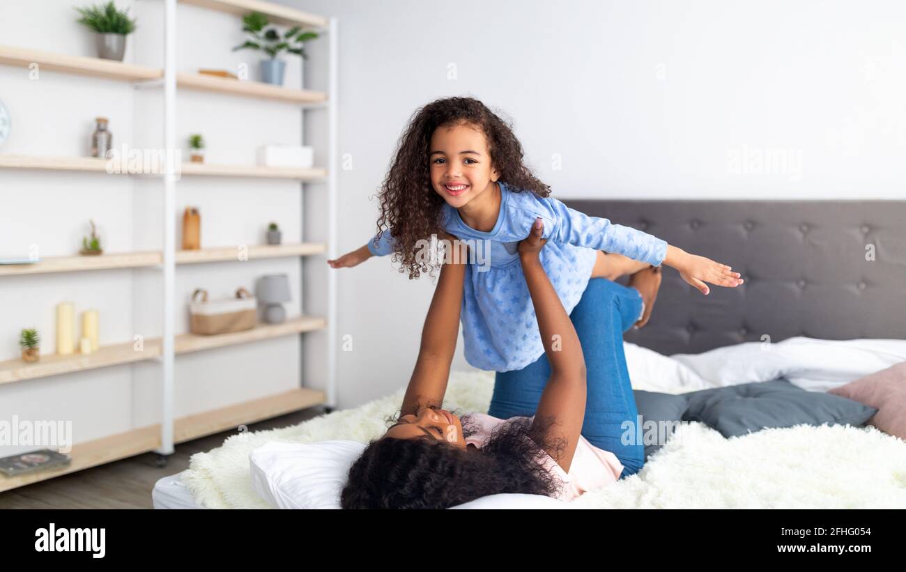 Glückliche schwarze Mutter und ihre süße Tochter spielen Flugzeug-Spiel auf dem Bett zu Hause, Panorama. Familienbindungszeiten Stockfoto