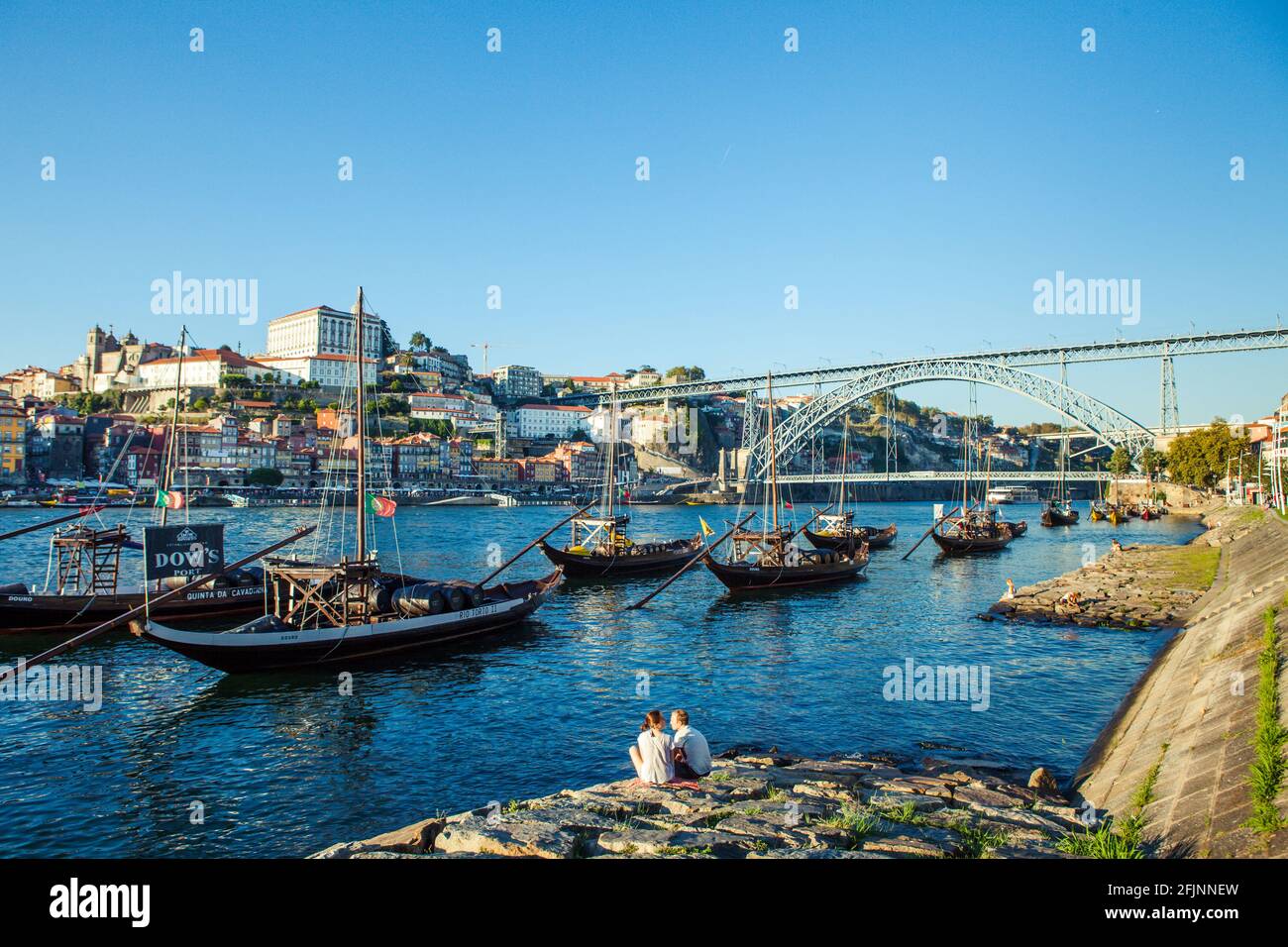 Ein Paar, das am Fluss Douro in Porto sitzt, mit den traditionellen Port Wines Barges und der Dom Luis Brücke im Hintergrund, Portugal. Stockfoto