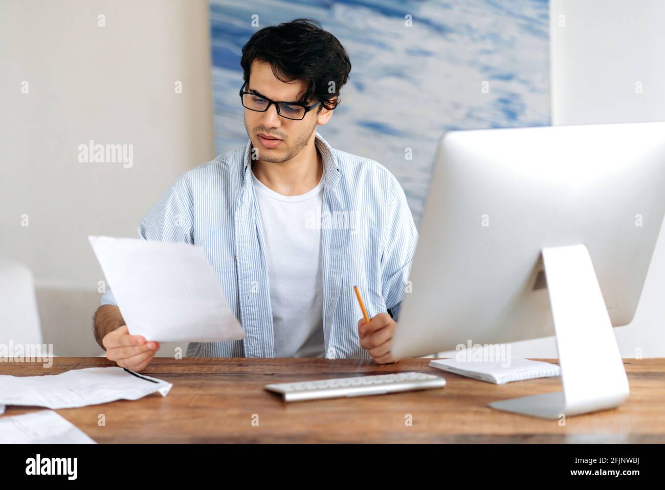 Konzentrierter selbstbewusster erfolgreicher gutaussehender hispanischer Mann mit Brille, Freiberufler, Student oder Geschäftsmann, der am Schreibtisch mit Computer arbeitet oder studiert und Geschäftsdokumente durchschaut Stockfoto