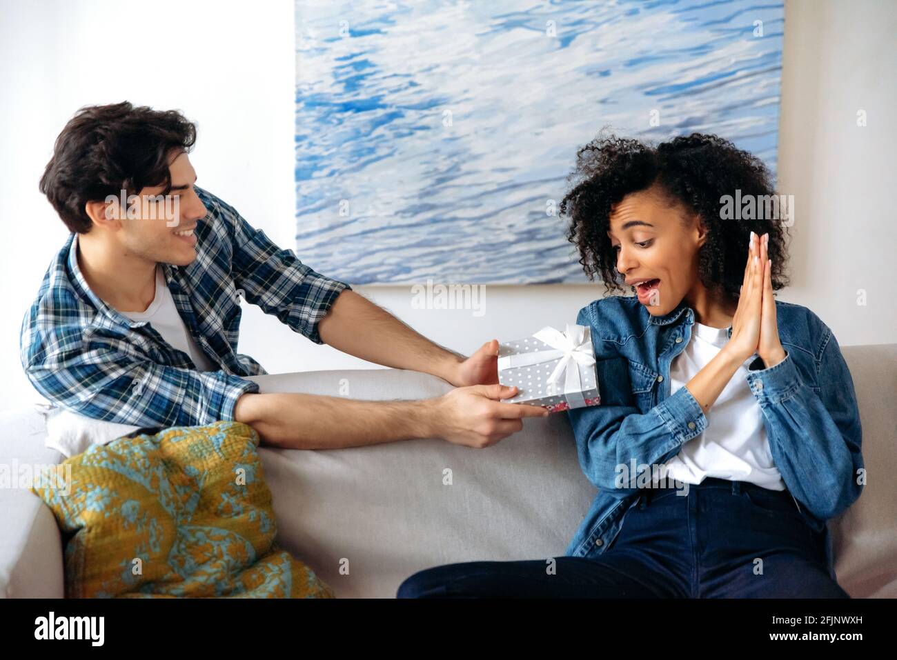 Glücklich überrascht junge attraktive afroamerikanische Frau erhält eine schöne Geschenkbox von ihrem fürsorglichen Latino-Freund zu einem Geburtstag oder Beziehungsjubiläum, schaut auf das Geschenk, klatscht in die Hände Stockfoto