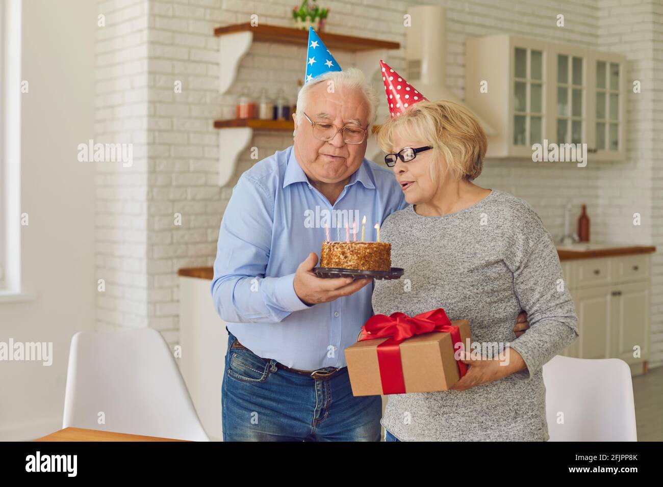 Glückliches reifes Paar in kegelförmigen Hüten, das Kerzen auf einem Kuchen ausbläst. Stockfoto