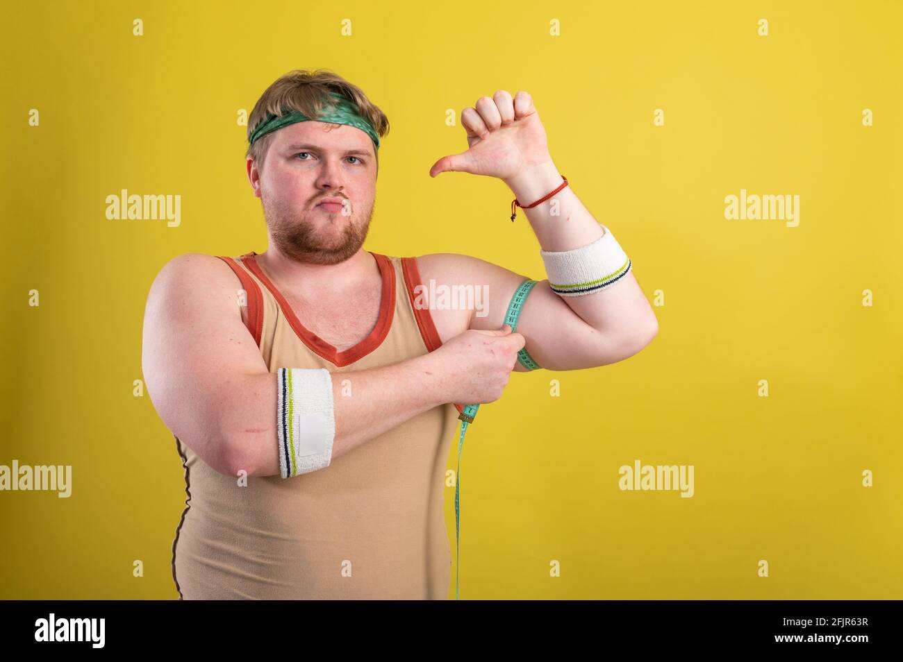 Ein lustiger, fröhlicher, fetter Mann in Sportkleidung mißt seine Arme. Übergewichtiger Mann geht in den Sport. Gelber Hintergrund isoliert Stockfoto