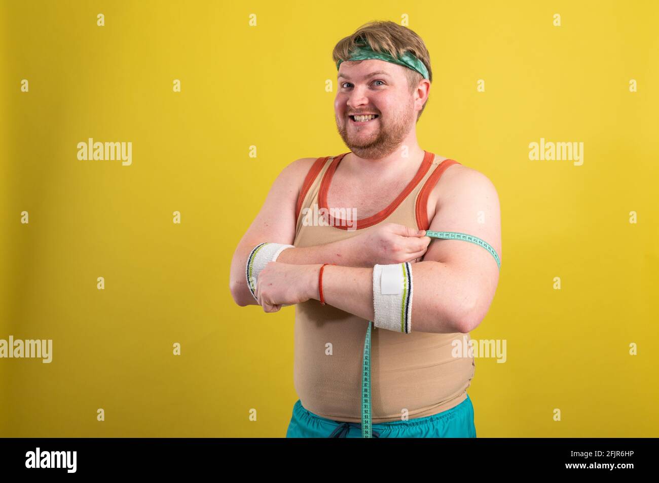 Ein lustiger, fröhlicher, fetter Mann in Sportkleidung mißt seine Arme. Übergewichtiger Mann geht in den Sport. Gelber Hintergrund isoliert Stockfoto