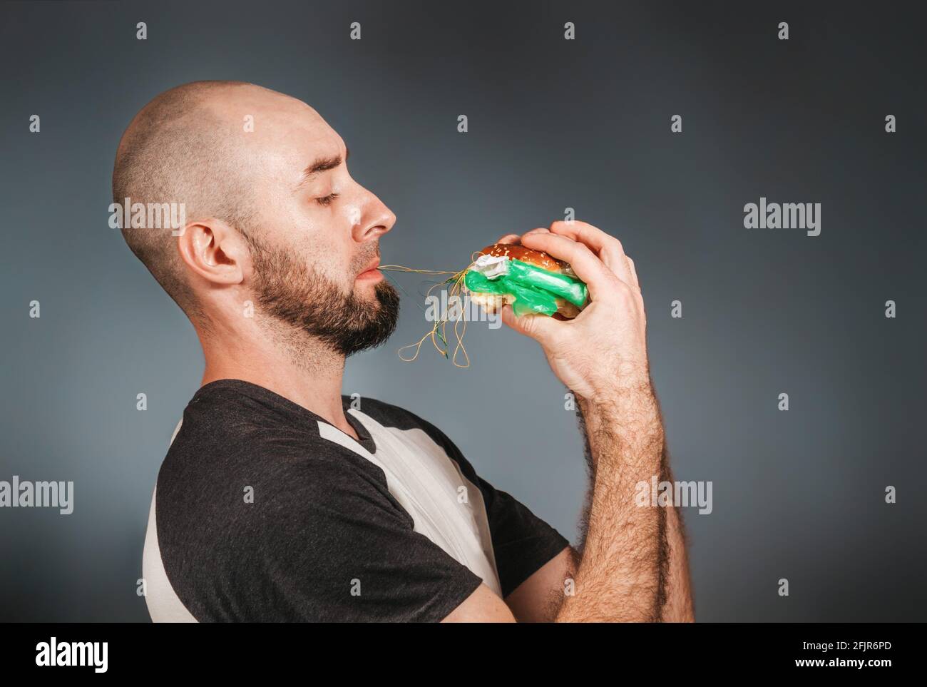 Umweltverschmutzung und Lebensmittelindustrie. Ein kahler Mann mit Bart hält mit beiden Händen einen Hamburger, aus dem sich eine Füllung aus Faden und Schutt zieht Stockfoto