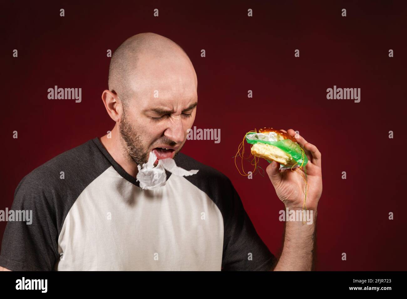 Das Konzept von Fast Food und Ökologie. Ein kahler Mann mit Bart, einem Hamburger haltend und einer Füllung aus Plastik. Burgunder Hintergrund. Stockfoto