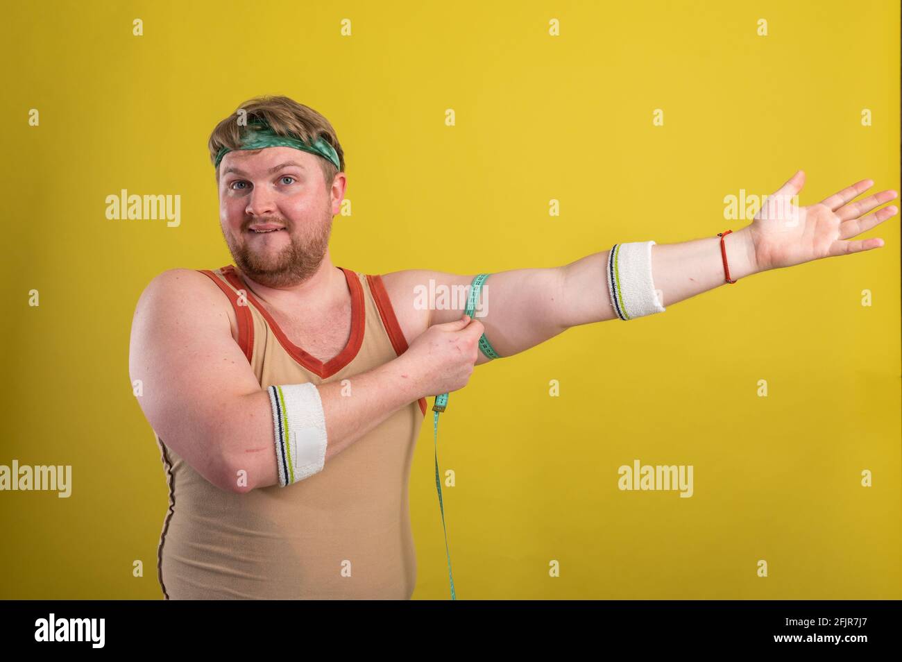 Ein lustiger, fröhlicher, fetter Mann in Sportkleidung mißt seine Arme. Übergewichtiger Mann geht in den Sport. Gelber Hintergrund isoliert Stockfoto
