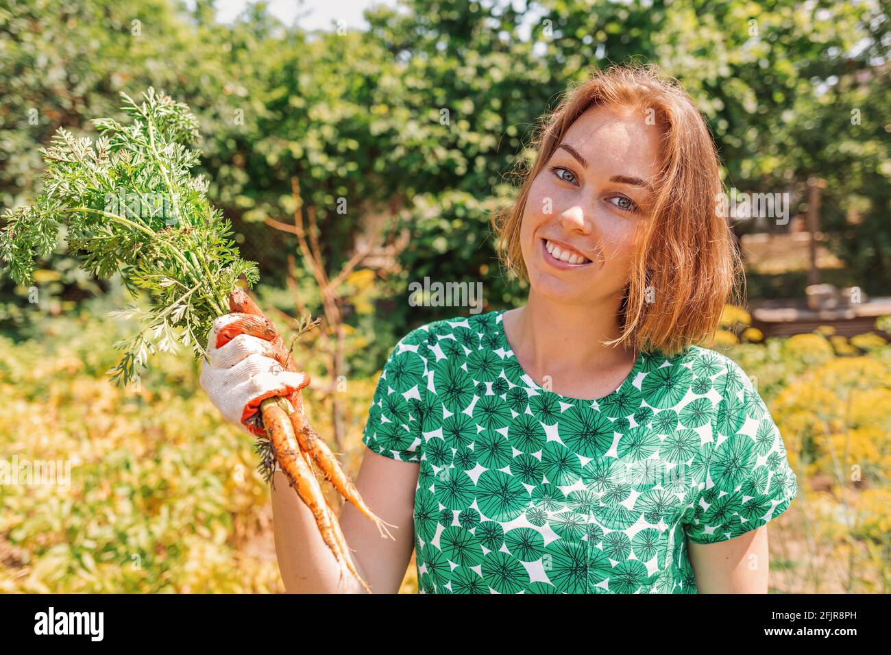 Eine kaukasische junge Frau lächelt und hält ein paar frisch gepflückte Karotten. Vegetation im Hintergrund.Konzept der Ernte und Gartenarbeit. Stockfoto