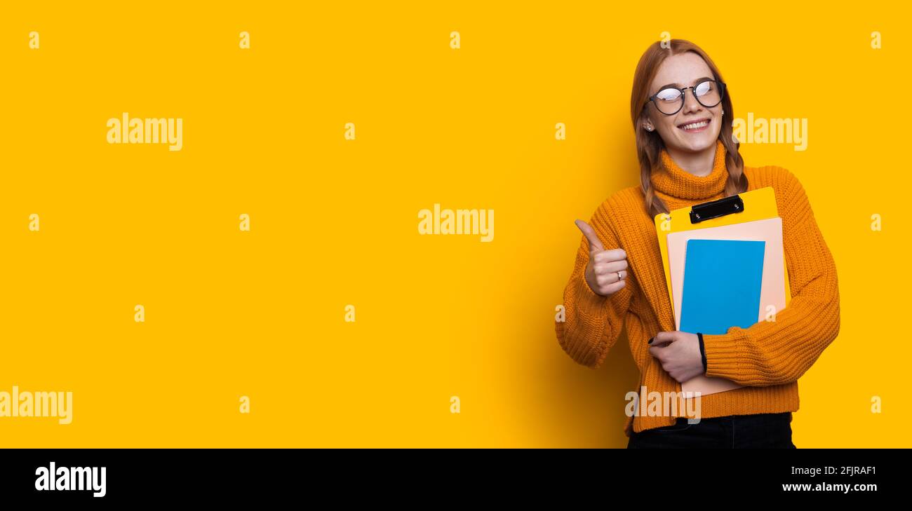 Der rothaarige Student stikelt das okay-Zeichen auf dem er posiert Eine gelbe Studiowand mit freiem Platz, an der einige Ordner befestigt sind Stockfoto