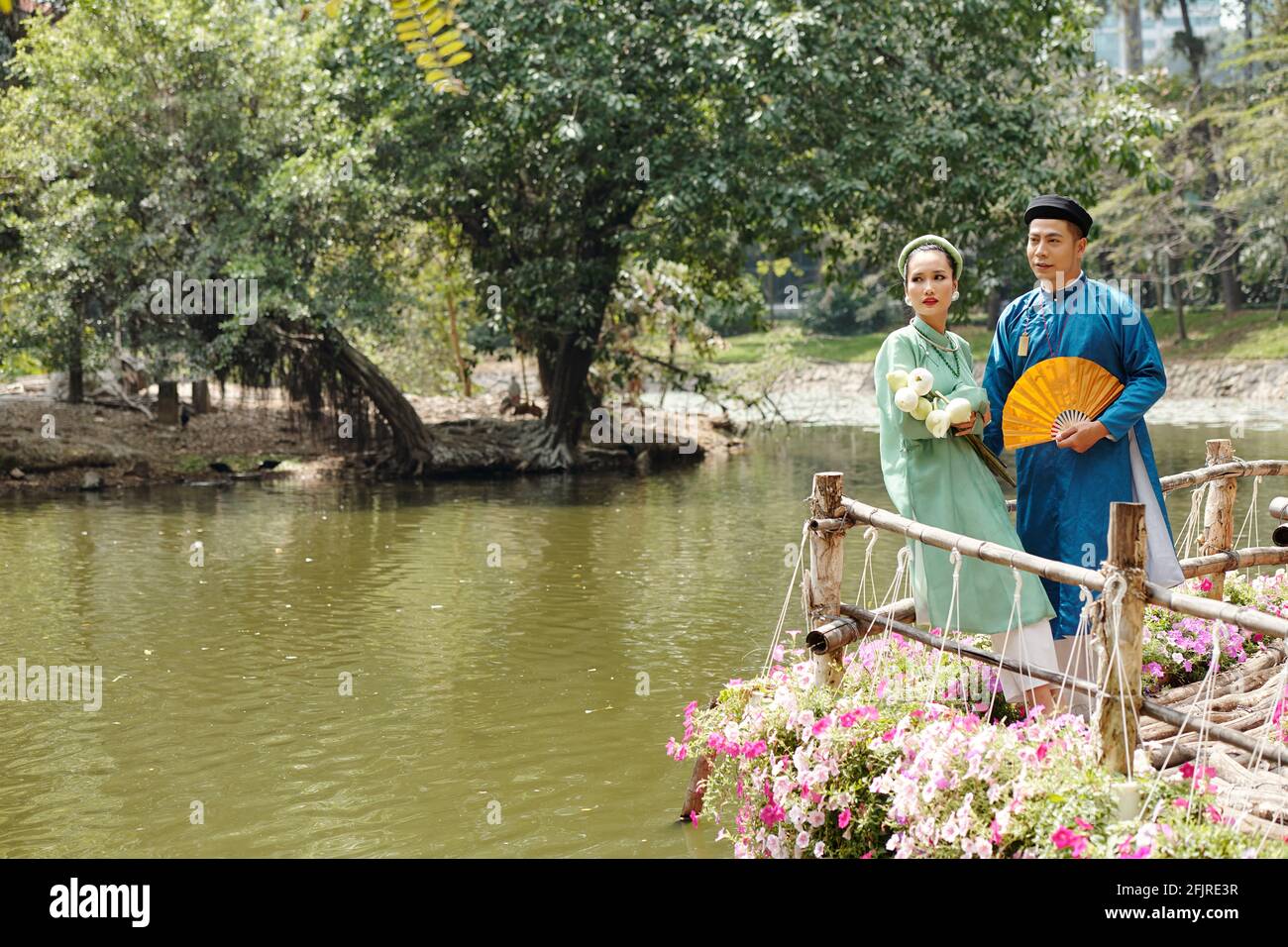 Vietnamesische Braut und Bräutigam in traditionellen Ao dai Kleidern und Kopfbedeckung namens khan dong, die am Teich im Stadtpark steht Stockfoto