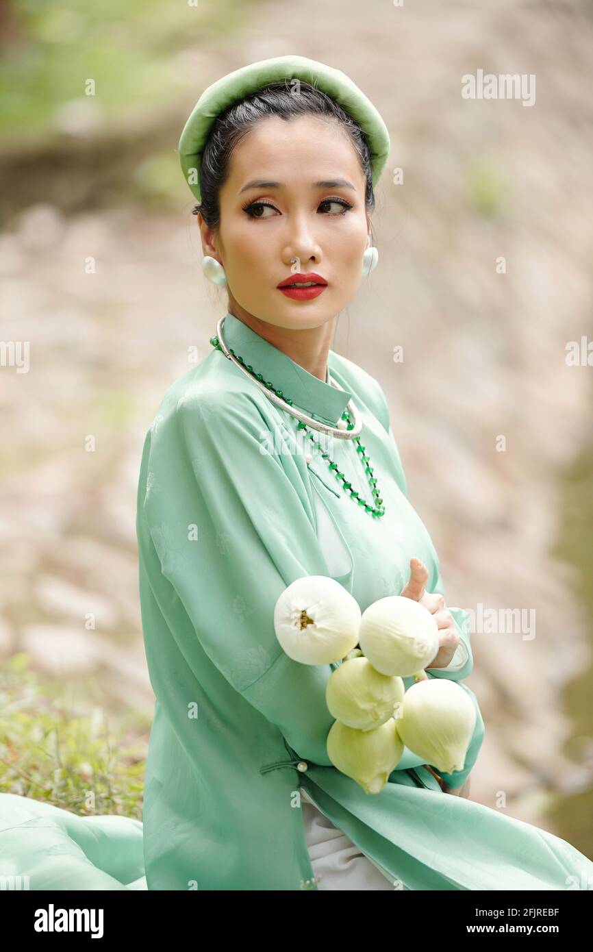 Porträt einer jungen ernsthaften Frau in hellem Minzkleid und Kopfbedeckung im Freien mit einem Haufen Lotusblumen in den Händen Stockfoto