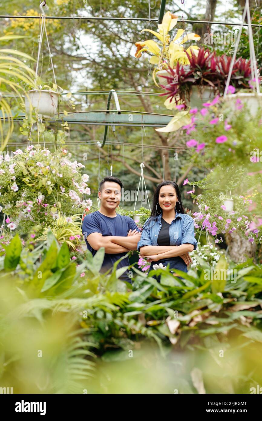 Glückliche junge asiatische Gewächshausbesitzer stehen im Gewächshaus mit Pflanzen Und Blumen in der Umgebung Stockfoto