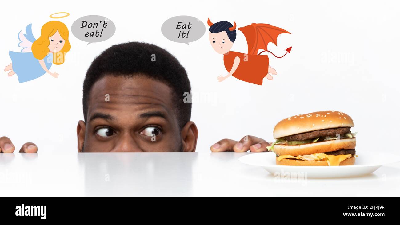 Hungriger afroamerikanischer Kerl, der ungesunde Burger auf dem Tisch anschaut, ihn zu essen sucht, von Teufel und Engel versucht wird Stockfoto