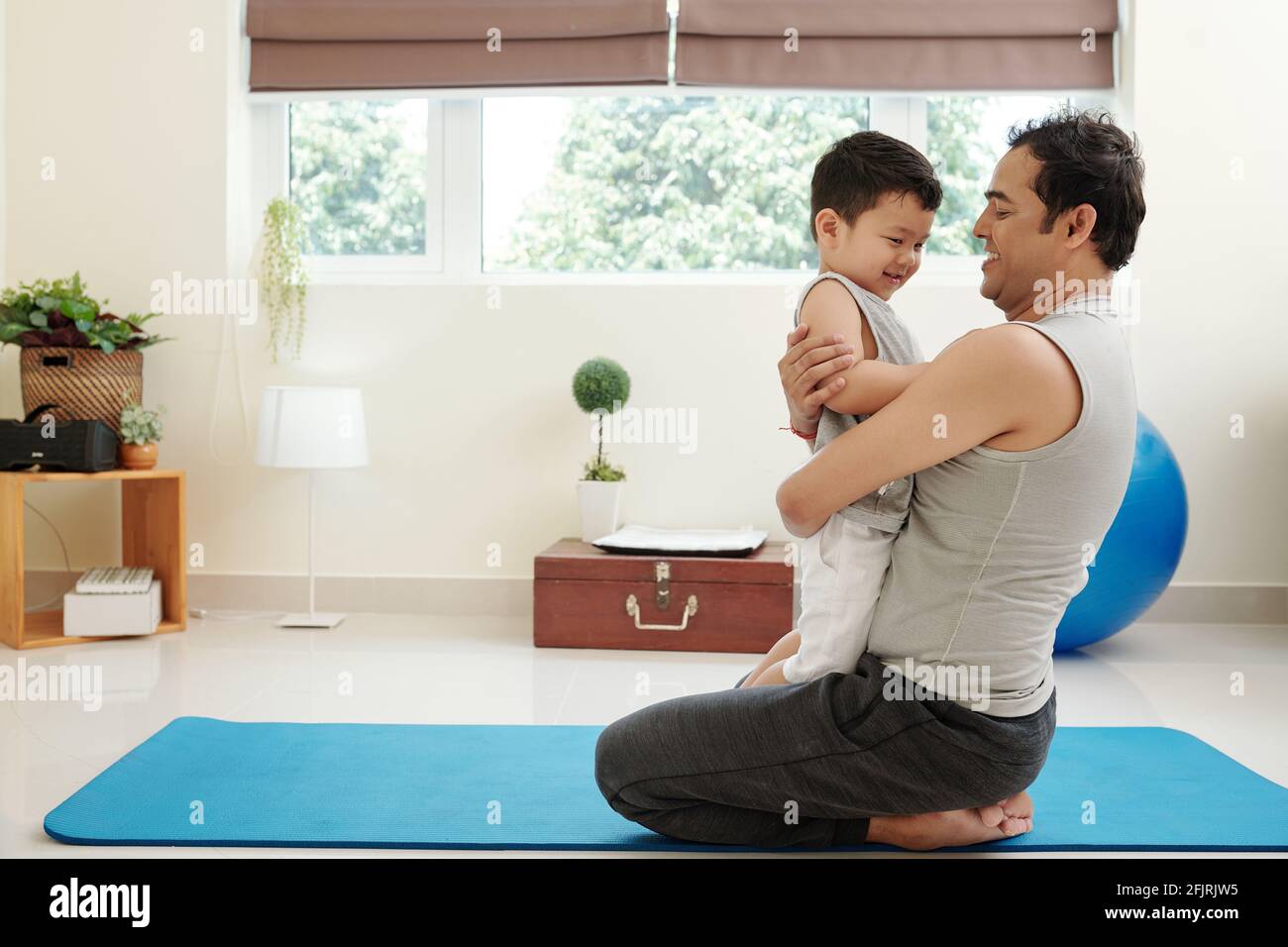 Glücklicher Vater, der auf einer Yogamatte sitzt und sein Lächeln umarmt Kleiner Sohn Stockfoto