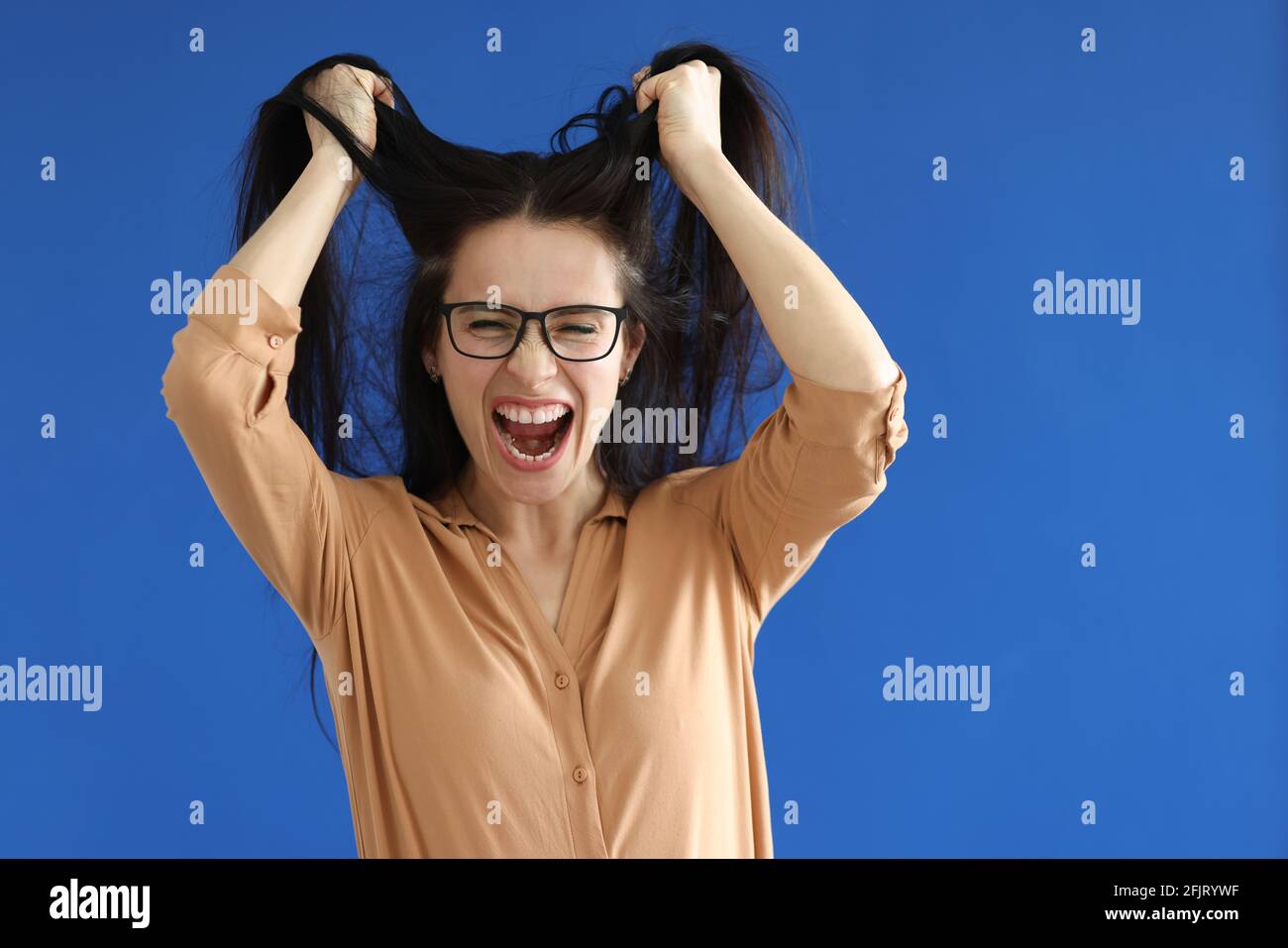 Wütende Frau mit Brille, die ihr Haar auf blauem Hintergrund zerreißt Stockfoto