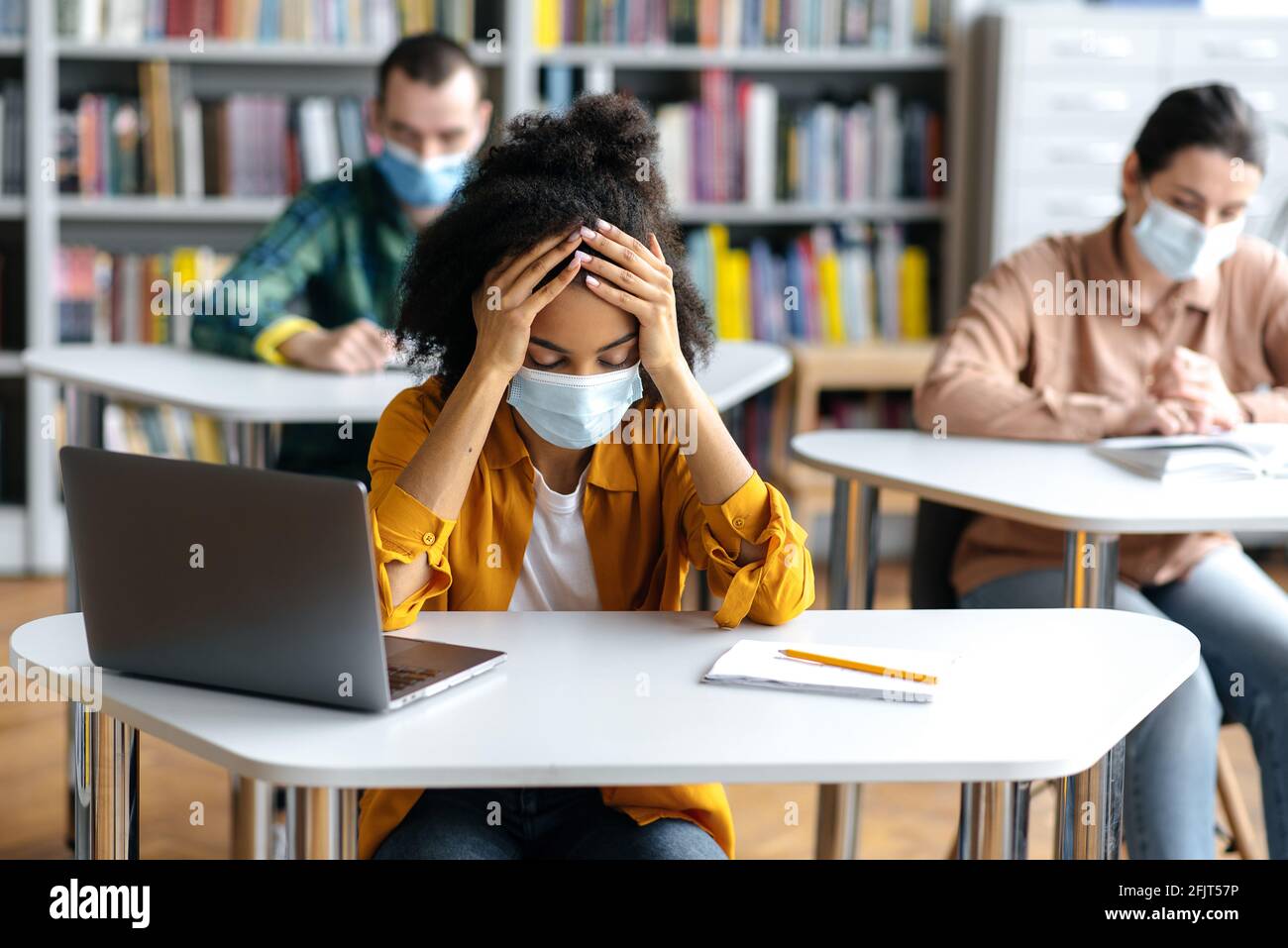 Aufklärung während einer Pandemie. Frustriertes, überarbeitetes afroamerikanisches Studentenmädchen mit Schutzmaske sitzt an einem Tisch in der Bibliothek, bereitet sich auf Prüfungen vor, müde, Klassenkameraden sitzen im Hintergrund Stockfoto