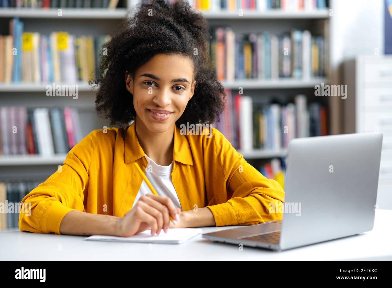 Porträt niedlich erfolgreiche afroamerikanische Studentin sitzt am Tisch in der Universitätsbibliothek, trägt stilvolle Kleidung, mit Laptop, macht Hausaufgaben oder bereitet sich auf die Prüfung vor, schaut auf die Kamera, lächelt Stockfoto