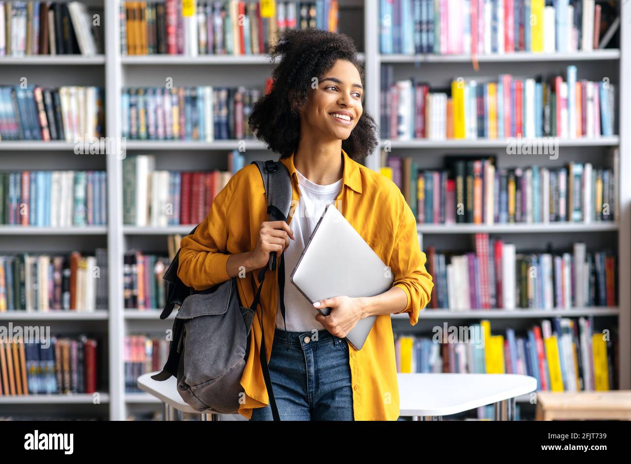 Porträt einer fröhlichen, hübschen afroamerikanischen Studentin, die vor dem Hintergrund von Bücherregalen in der Universitätsbibliothek steht und Laptop und Rucksack in der Hand hält, die zur Seite schauen und angenehm lächeln Stockfoto