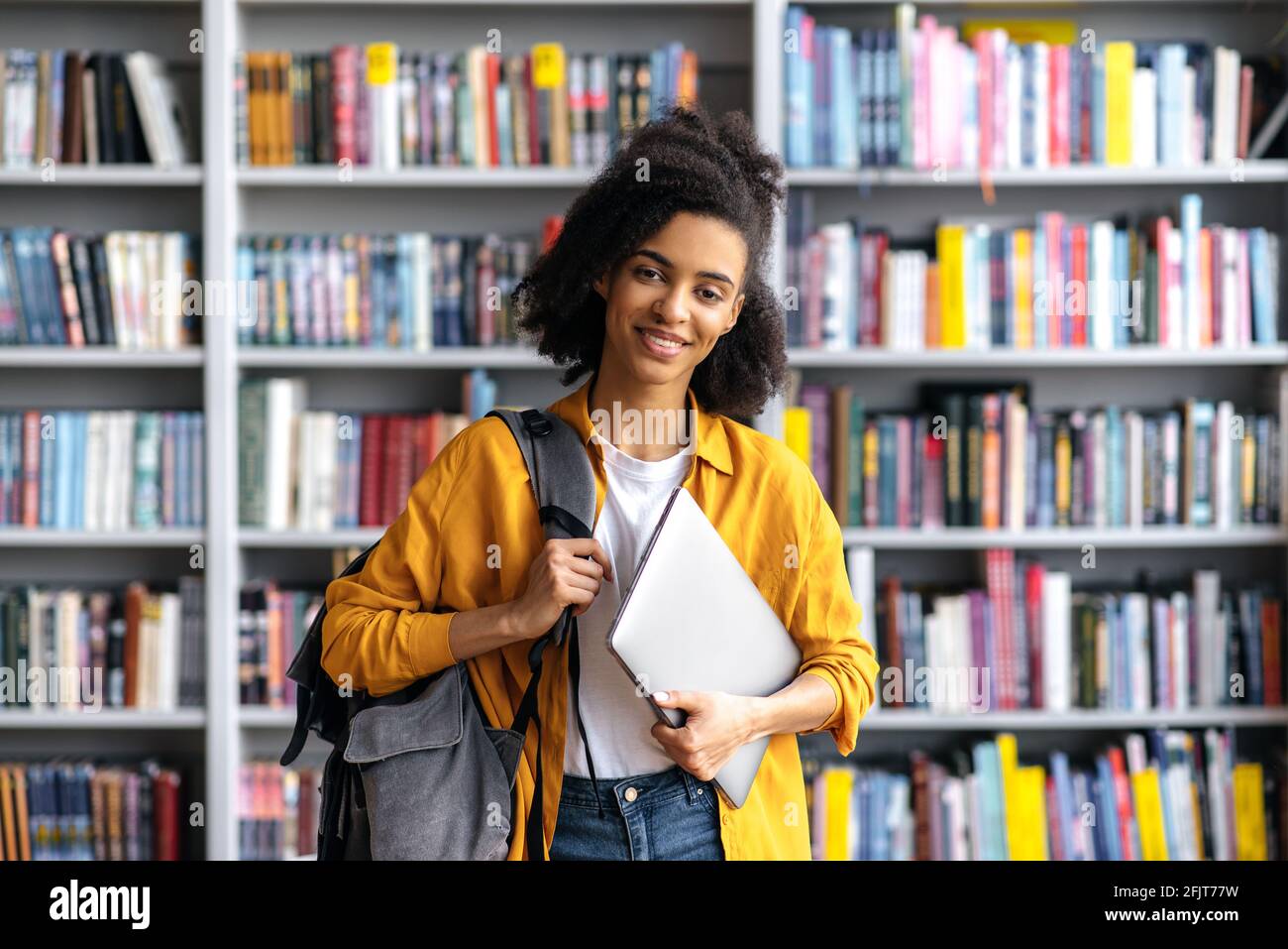 Portrait selbstbewusst glücklich Attraktive afroamerikanische Studentin im Teenageralter, die vor dem Hintergrund von Bücherregalen in der Universitätsbibliothek steht, Laptop und Rucksack in der Hand hält, auf die Kamera schaut und freundlich lächelt Stockfoto