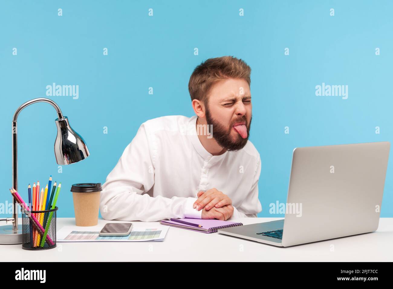 Lustiger unzufriedener Mann mit Bart, der die Zunge zeigt, sprechender Videoanruf auf dem Laptop, der am Arbeitsplatz sitzt, Spaß hat und kindhaftes Verhalten demonstriert. Ind Stockfoto