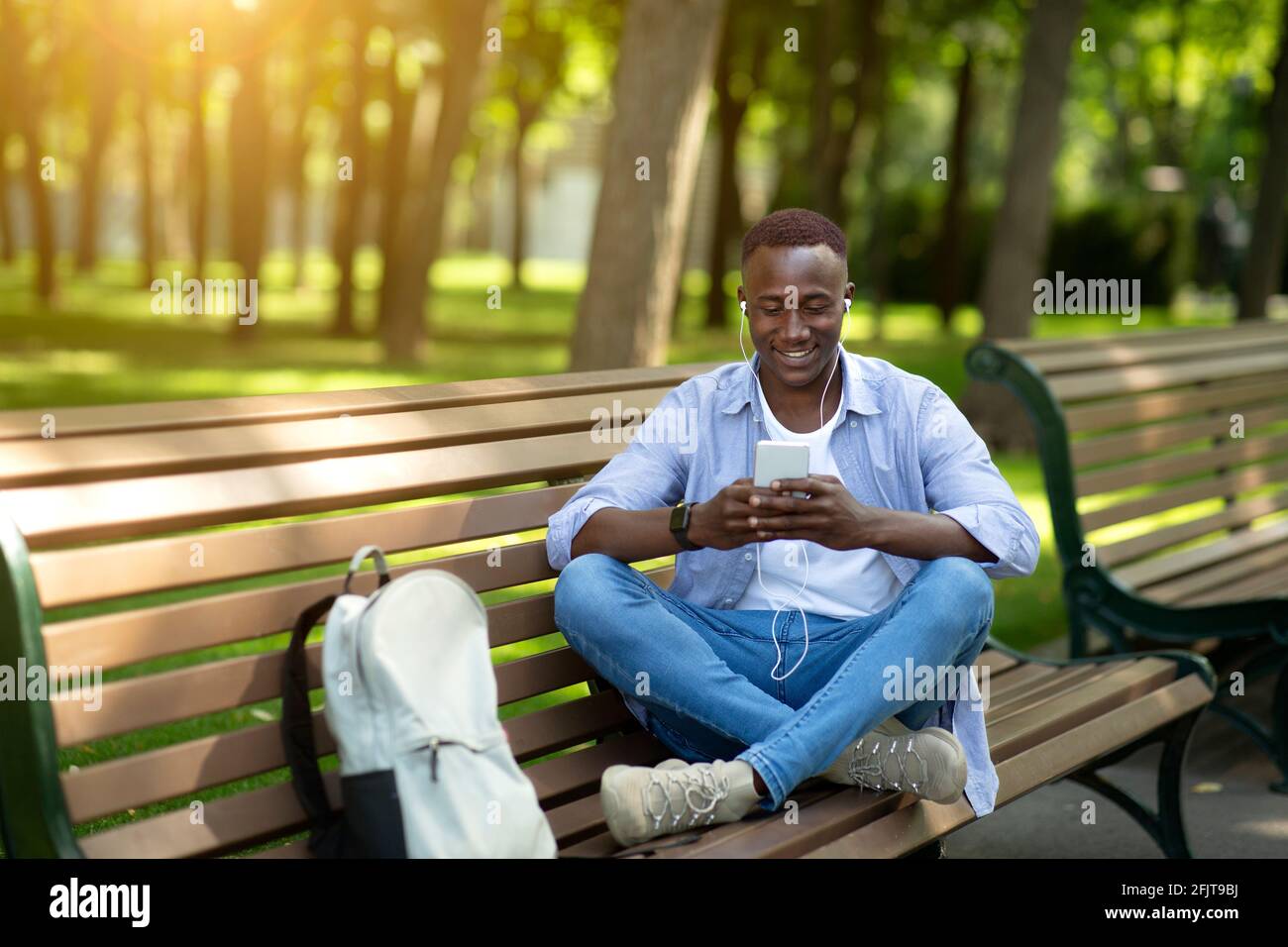 Funky schwarzer Kerl in Ohrhörern hört seine Lieblingsmusik online von Handy auf Bank im Park, Kopierer Platz Stockfoto