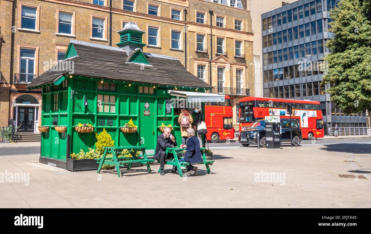 Londoner Cabmen's Shelter. Ein historisches Gebäude, das traditionell Taxifahrern Zuflucht bietet. Ein modernes Elektrotaxi lädt im Hintergrund. Stockfoto