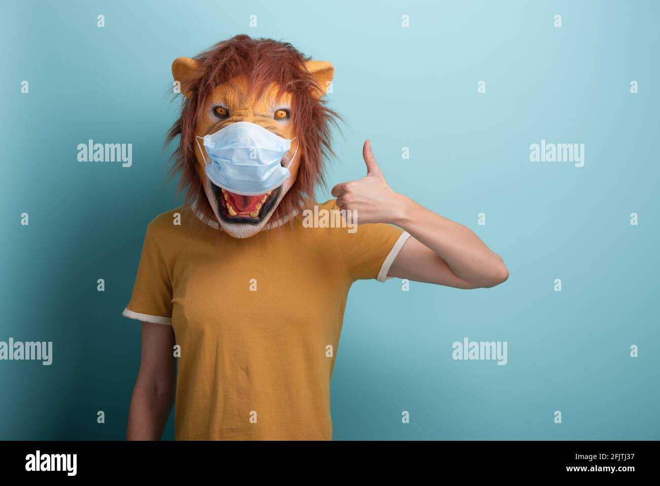 Junge Frau trägt Löwen und Schutz medizinische Maske zeigen Daumen nach oben Zustimmung Geste, isoliert auf blauem Hintergrund. Stockfoto