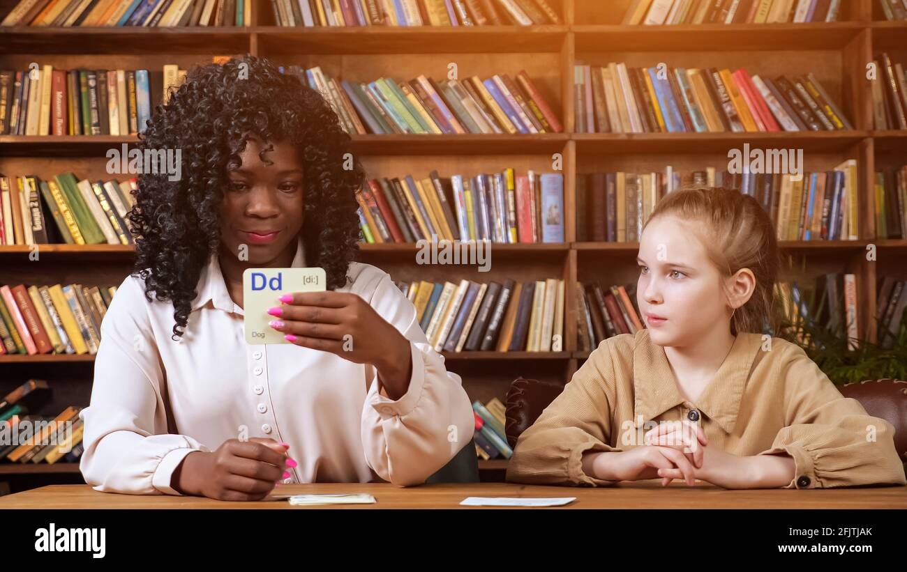 Professionelle schwarze Lehrerin wiederholt Briefe mit einer jungen Dame, die Karteikarten gegen braune Holzregale zeigt, mit Büchern zu Hause, covid Lockdown, Sonnenlicht Stockfoto