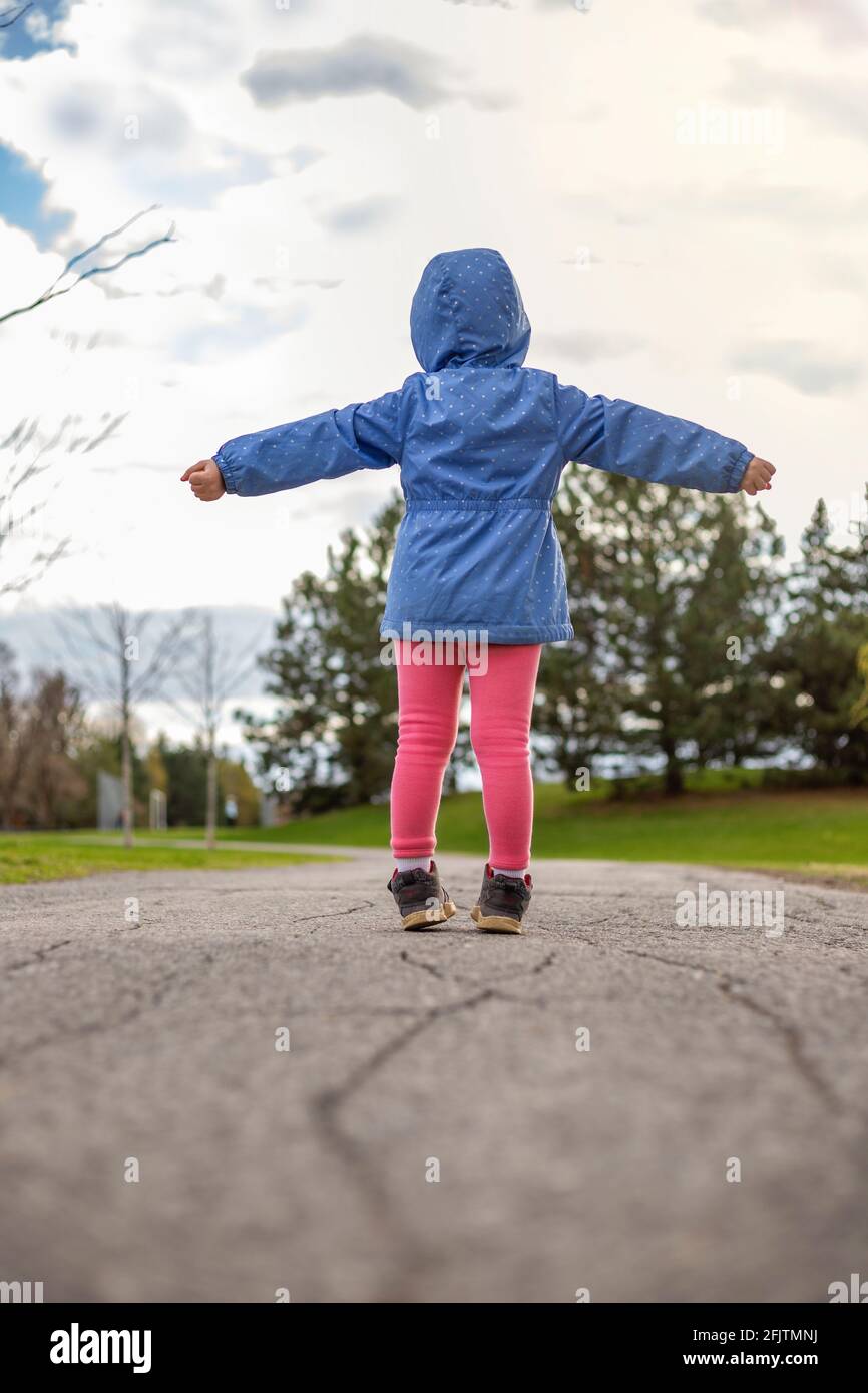 Kleines Mädchen, das im Frühjahr auf einer Straße im Park steht. Kind von hinten genießt an einem warmen, sonnigen Tag den Spaziergang und hält die Hände an den Seiten hoch. Stockfoto