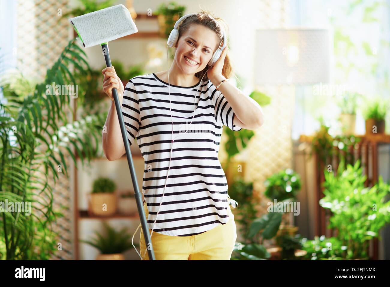 Lächelnde, moderne Hausfrau in gestreiftem Hemd mit Mopp und Kopfhörern, die an sonnigen Tagen im modernen Haus Hausarbeit leistet. Stockfoto