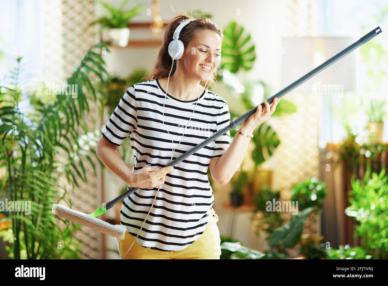 Glückliche Hausfrau mittleren Alters in gestreiftem Hemd mit Mopp und Kopfhörern, die an sonnigen Tagen zu Hause Hausarbeit leistet. Stockfoto