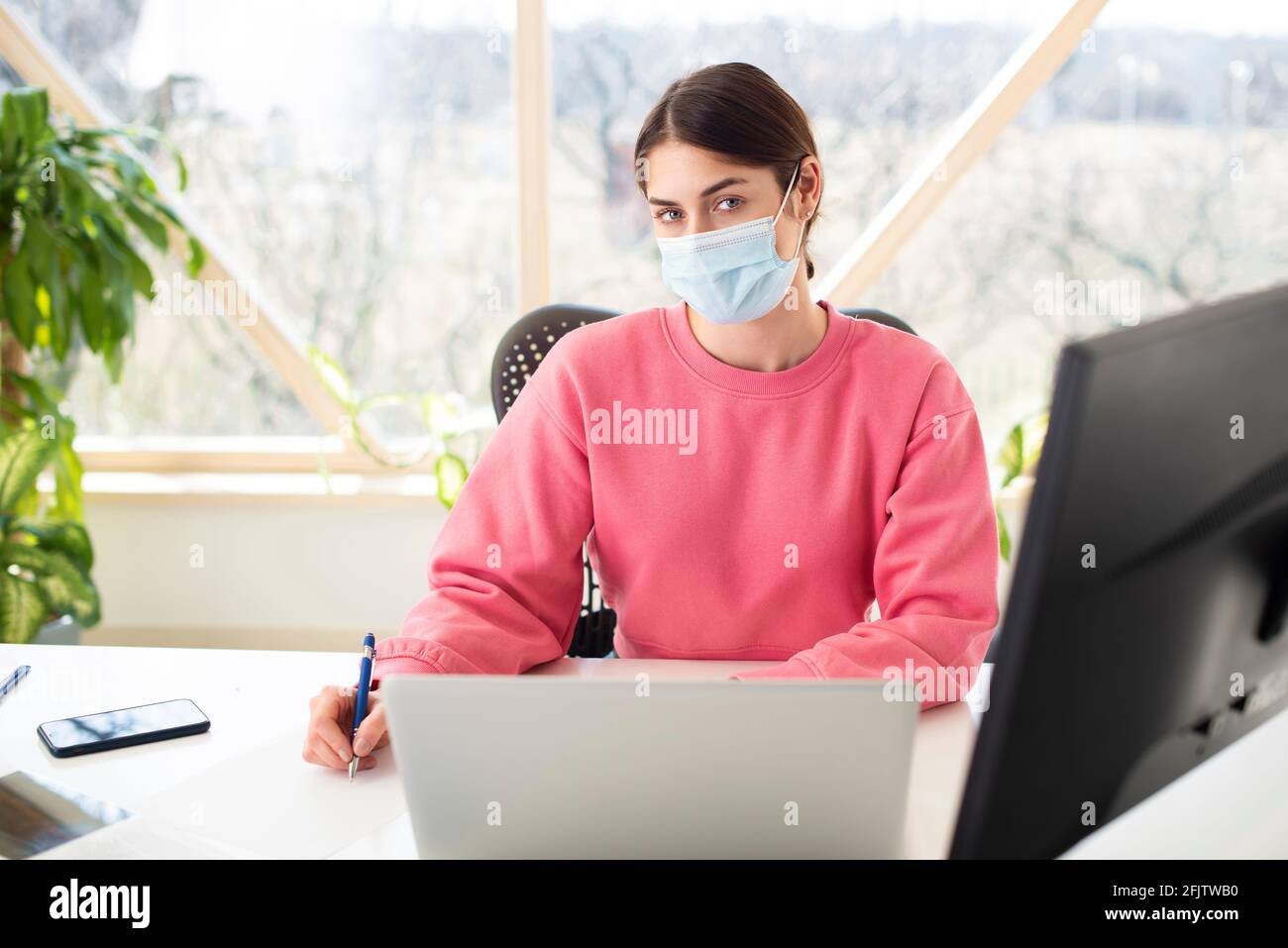 Aufnahme einer schönen Frau, die eine Gesichtsmaske trägt, während sie hinter ihrem Computer sitzt und arbeitet. Heimbüro. Stockfoto