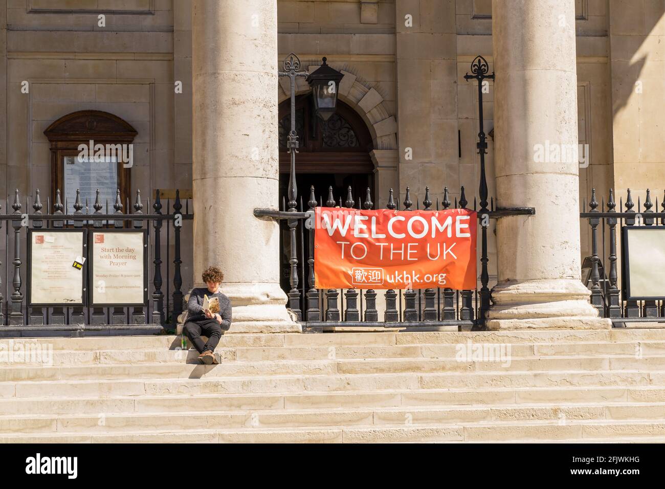 Mann, der in der Sonne sitzt und vor ihm ein Buch liest Eines „Welcome to the UK“-Zeichens auf den Schritten St. Martin in der Fields-Kirche am Charing Cross Road London Stockfoto