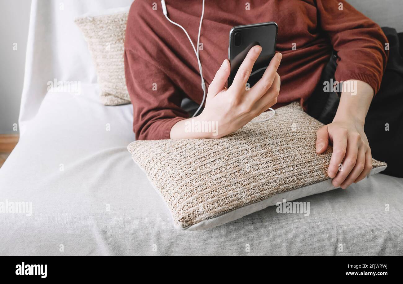 Die Hände der Frau ruhen auf den Kissen eines Sofagarnitur Ein Mobiltelefon, an das Kopfhörer zu Hause angeschlossen sind Stockfoto
