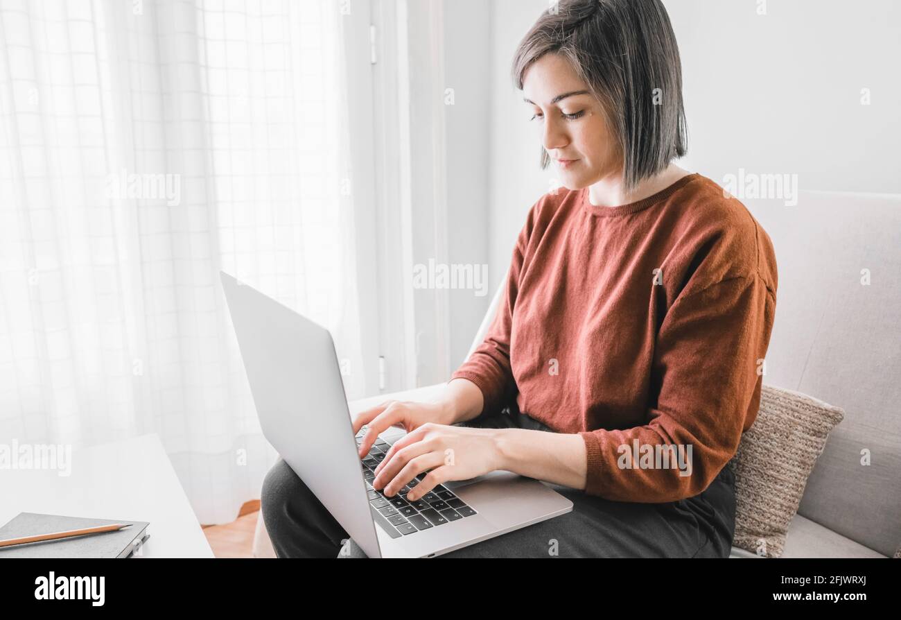 Frau, die in ihrem Laptop auf dem Sofa sitzt Ihr helles Wohnzimmer zu Hause mit einem grünen Notizbuch In einem weißen Tisch Stockfoto