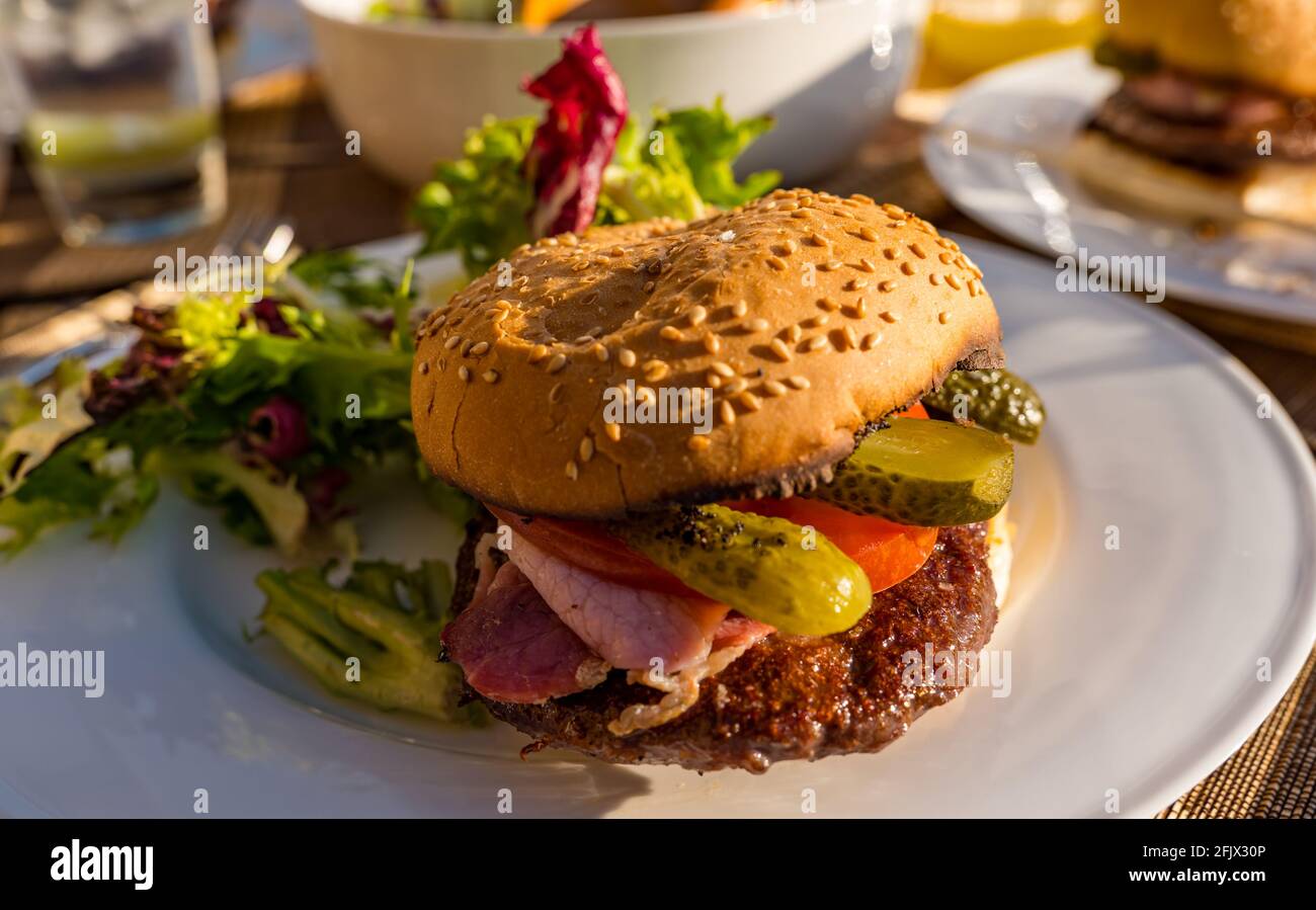 Rindfleisch-Burger mit Speck, Tomaten und Dill-Pickle auf einem weißen Teller mit Salat auf einem Tisch im Freien bei Sonnenschein Stockfoto