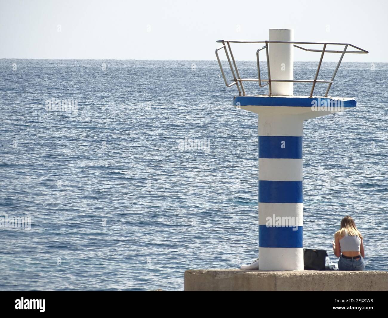 Junge Frau, die in der Nähe eines Leuchtturms sitzt, der vom Meer umgeben ist Unter dem Sonnenlicht Stockfoto