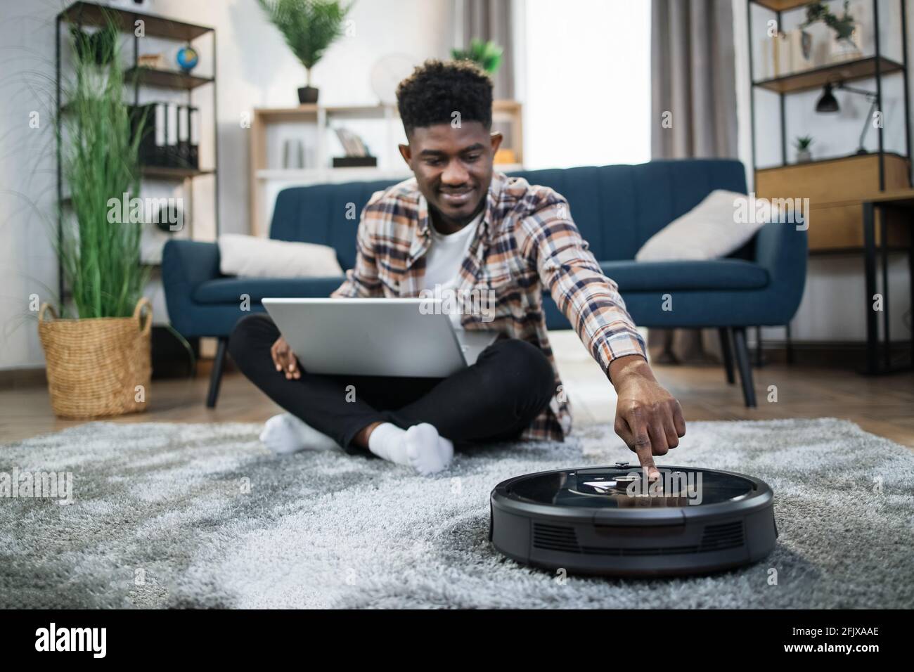 Lächelnder afrikanischer Mann, der Roboter-Staubsauger anmacht, während er  Teppich im Wohnzimmer putzt. Glücklicher Kerl, der während des Haushaltes  mit einem modernen Gerät am Laptop arbeitet Stockfotografie - Alamy